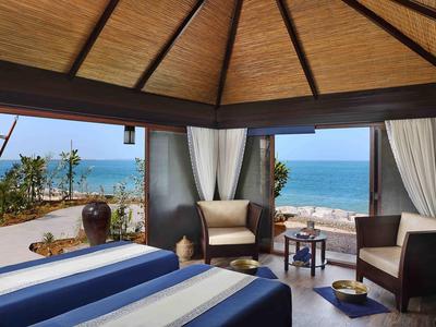 Offenes Hotelzimmer mit Blick auf das Meer, zwei Betten und Loungemöbeln unter Holzdach.