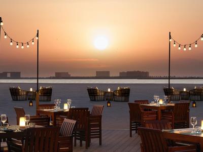 Ein Hotelsitzbereich am Wasser mit Tischen, Stühlen und Lichtern bei Sonnenuntergang.