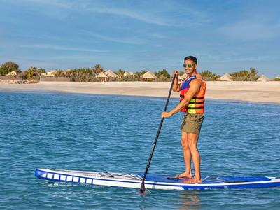 Person in orange Rettungsweste auf blauem Stand-up-Paddle-Board im Wasser vor Strand.