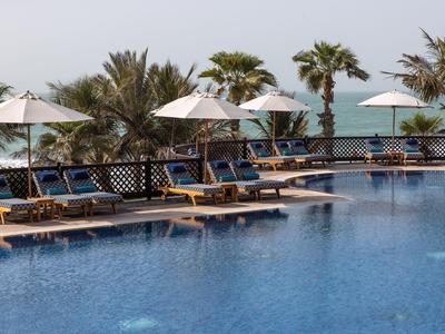Área de piscina con tumbonas, sombrillas y palmeras frente al mar en el fondo
