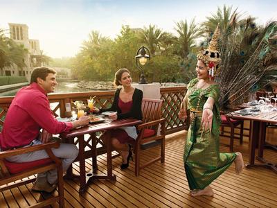 Una pareja está sentada en una terraza mirando a una mujer con vestido tradicional y plumas.
