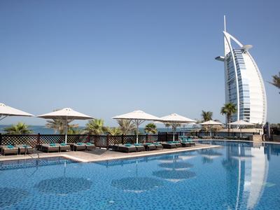 Moderní bazén s deštníky a hotelem Burj Al Arab v pozadí za jasné oblohy.