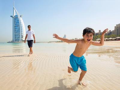 Chlapec běží po pláži s mužem a Burj Al Arab v pozadí.