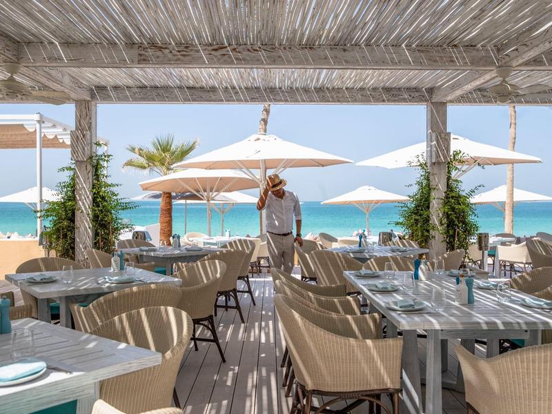 Otevřená plážová restaurace s prostřenými stoly, židlemi a výhledem na moře a slunečníky.