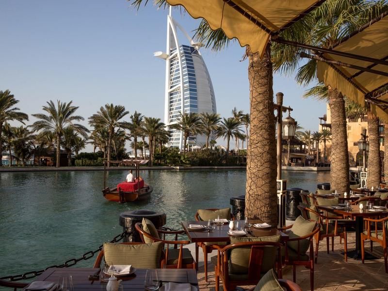 Venkovní restaurace s výhledem na vodu a Burj Al Arab v Dubaji za jasné oblohy.