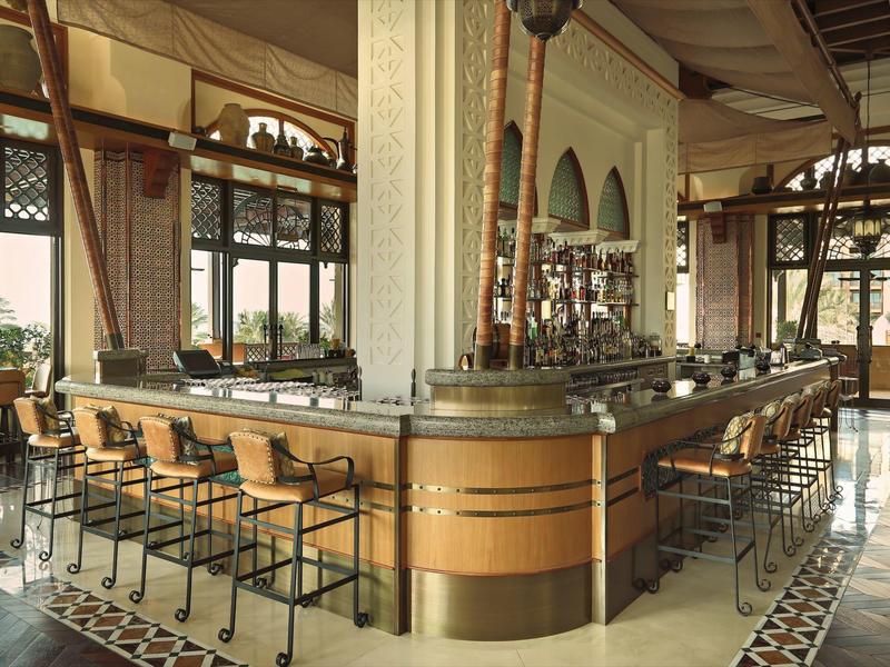 Elegantní hotelový bar s dřevěným pultem, stoličkami a velkými okny s výhledem ven.