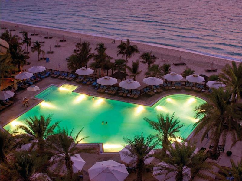 Osvětlený bazén vedle pláže s palmami a slunečníky při západu slunce.