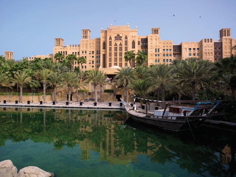 Tradiční loď před luxusním hotelem s palmami a modrou oblohou.