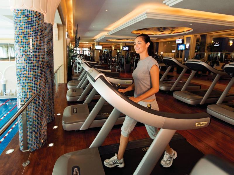 Žena cvičí na běžeckém pásu v moderní posilovně s výhledem na bazén.