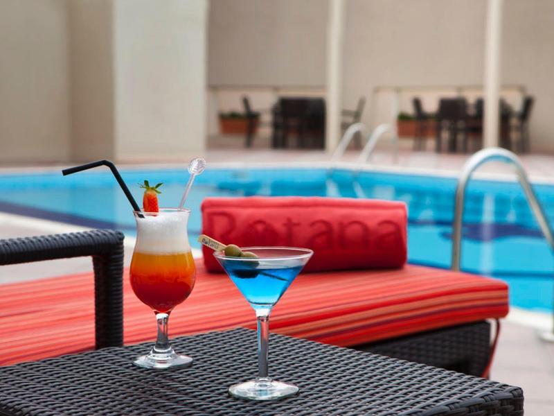 Barevné koktejly na stole vedle bazénu s červenými polštáři v pozadí.