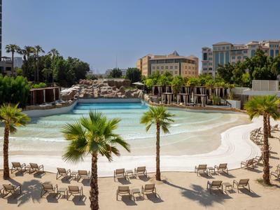 Velký hotelový bazén s lehátky, palmami a budovami v pozadí pod jasnou oblohou.