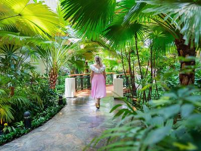 Žena v růžových šatech kráčí po zakřivené cestě tropickou hotelovou zahradou.