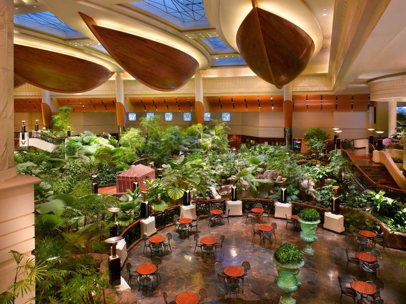 Velká hotelová hala s bujnou zelení, spoustou stolů a židlí pod moderním stropem.