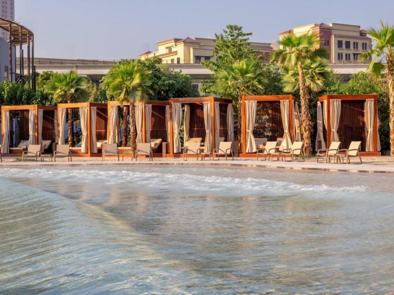 Pláž s dřevěnými kabinami a lehátky před vícepodlažními budovami při východu slunce.