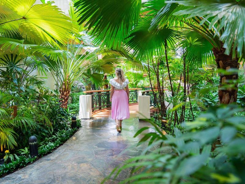 Žena v růžových šatech kráčí po zakřivené cestě tropickou hotelovou zahradou.