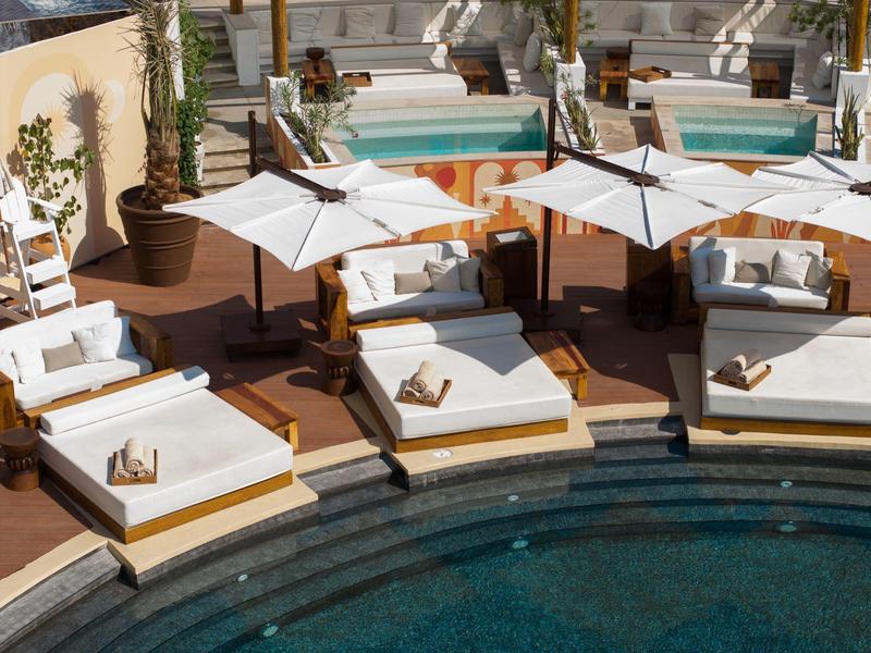 Several cushioned lounge chairs with umbrellas by a pool surrounded by palm trees.