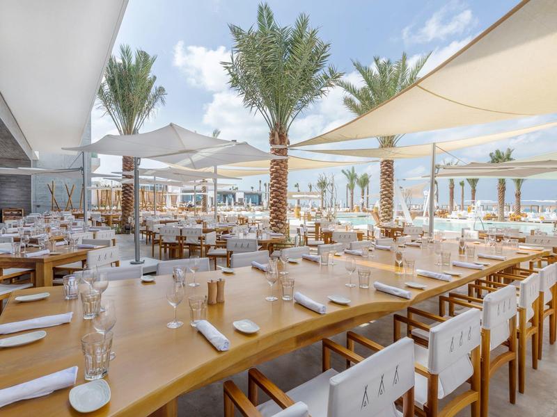 Modern restaurant with wooden tables and white chairs on the beach with palm trees and sun shades.