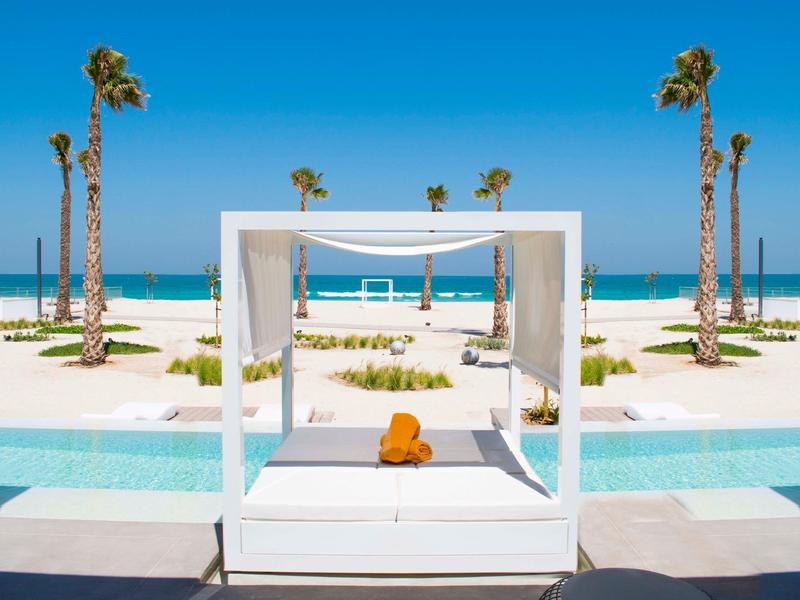 White daybed with cushions in front of beach, palm trees, and sea under blue sky.