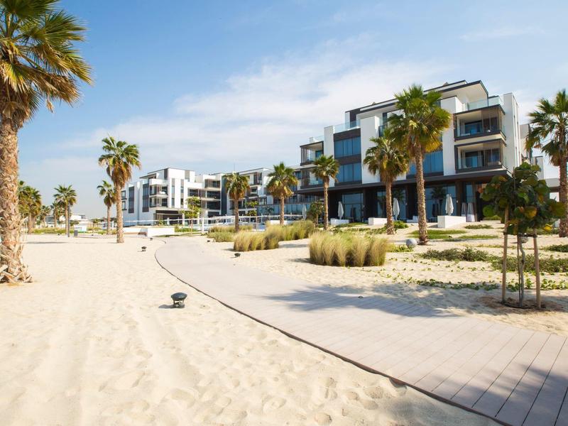 Modern apartments with palm trees on sandy beach and boardwalk under clear sky.