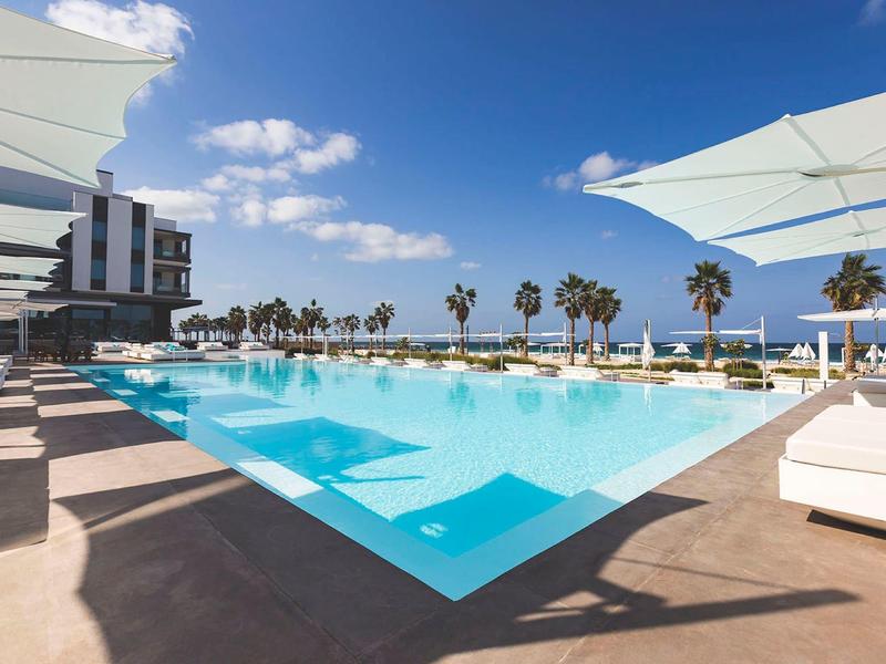 Large outdoor pool with sun loungers and umbrellas beside a hotel under a blue sky.
