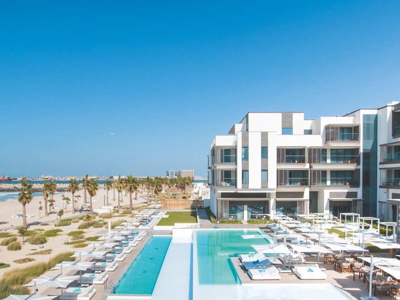 Modern hotel pool with sun loungers and palm trees under a clear blue sky.