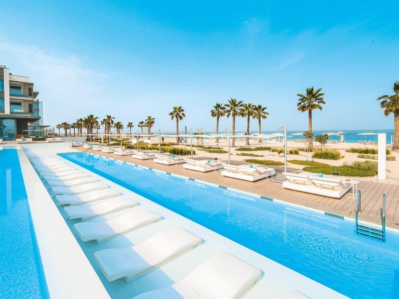 Modern hotel pool with built-in white loungers, palm garden, and beach view.