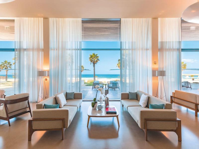 Bright hotel room with sofas, armchairs, and sea view in daylight.