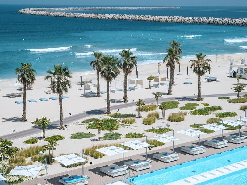 Beach with palm trees, loungers, and pool at a seaside resort.