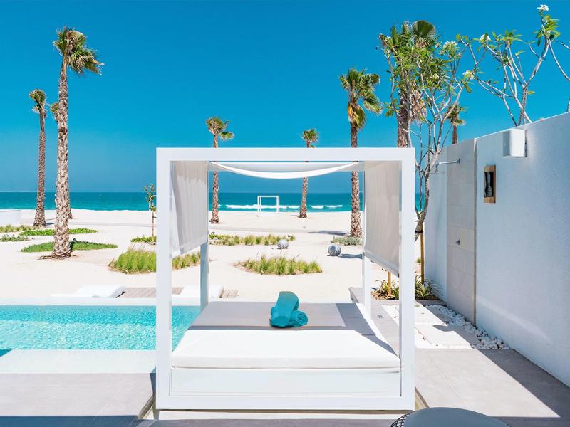 Modern white beach bed with pillows beside pool and palm trees by the sea.