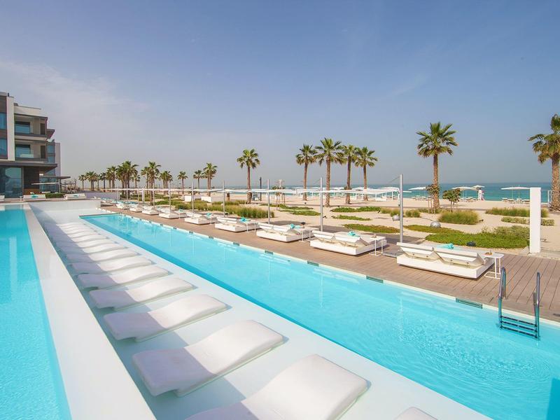Modern pool with white loungers by the beach lined with palm trees under clear sky.