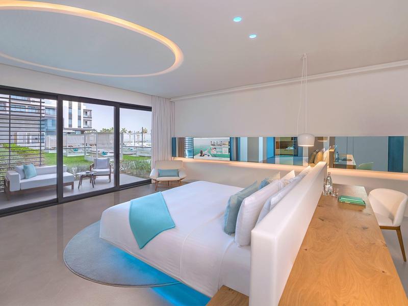 Modern hotel room with large bed, blue accents, and terrace access overlooking city and garden.