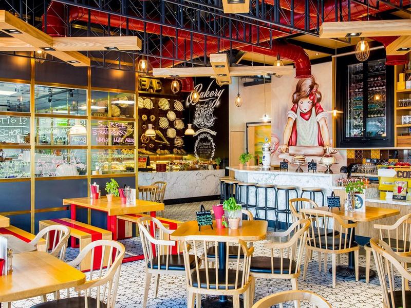 Kleurrijke eetruimte met houten stoelen, gele tafels en een chef-beeld achter de toonbank.