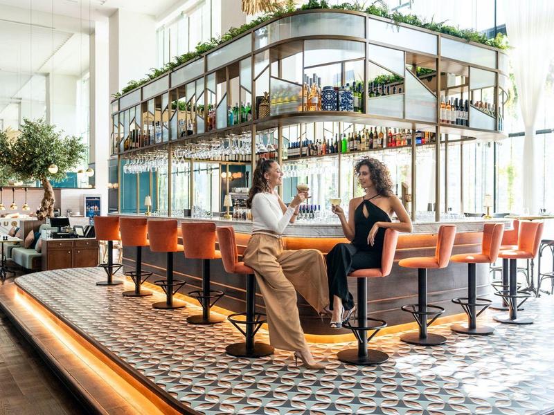 Twee personen praten bij een moderne hotelbar met oranje stoelen en decoratieve planten.
