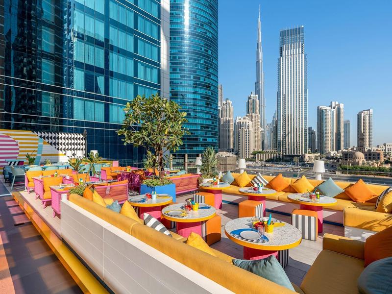 Kleurrijke dakterraslounge met kussens, tafels en stadsgezicht op de achtergrond.