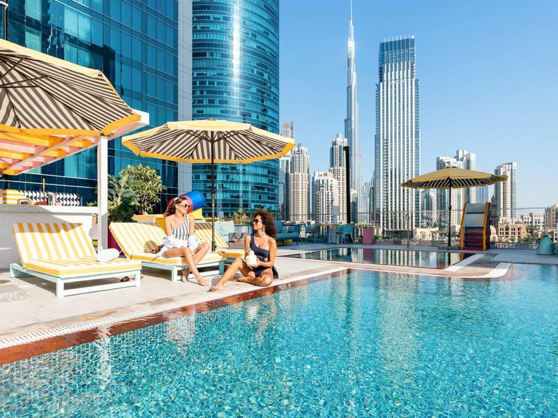 Twee vrouwen ontspannen bij een dakterraszwembad met ligstoelen en een stadsgezicht op de achtergrond.