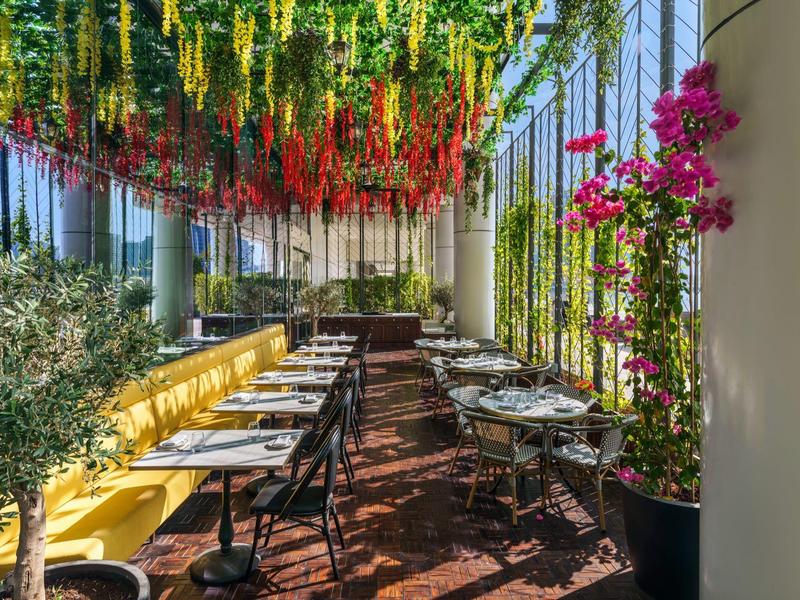 Buitenterras van restaurant met hangende bloemen en gele zitplaatsen onder glazen dak