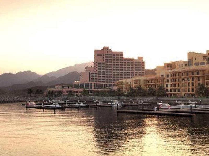 Hafen mit mehreren Booten vor Gebäuden und Bergen bei Sonnenuntergang.