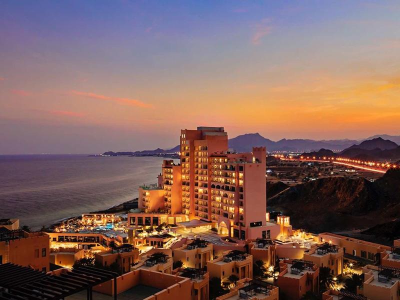 Stadt mit beleuchteten Gebäuden am Meer bei Sonnenuntergang, Hügel im Hintergrund, orange-blauer Himmel.