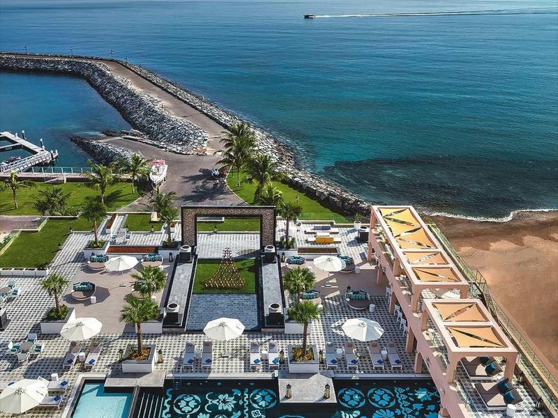 Blick auf Hotelanlage mit Pool, Palmen und Blick auf ruhiges Meer unter blauem Himmel.