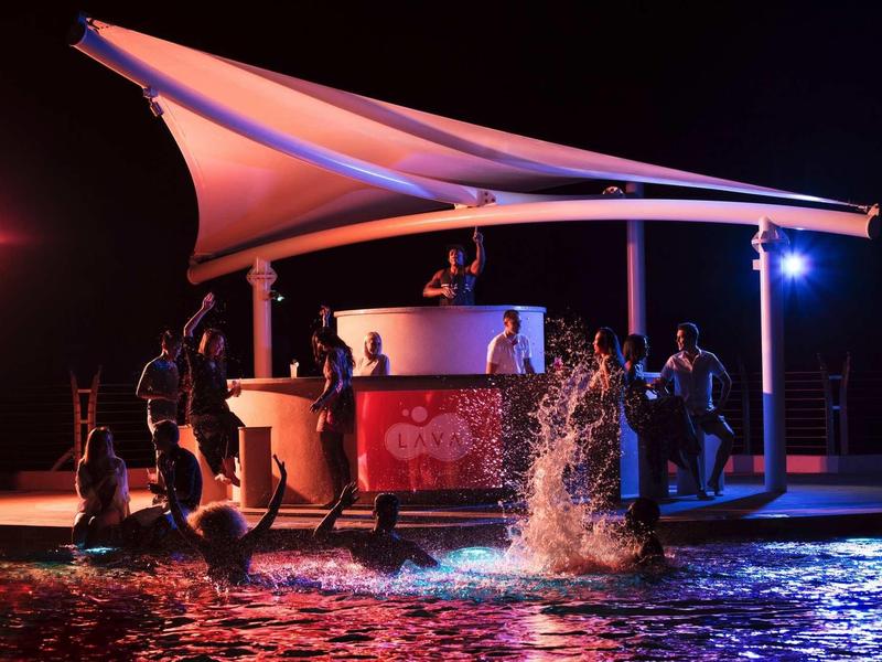 Menschen feiern bei Nacht an einem beleuchteten Strandpavillon mit Wasser und Spritzern im Vordergrund.