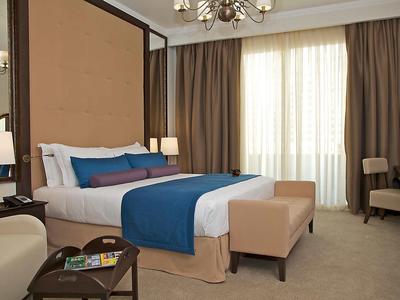 Elegantní hotelový pokoj s velkou postelí, modrými detaily, křesly a velkými okny se závěsy.