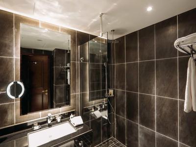 Moderní koupelna s tmavými dlaždicemi, osvětleným zrcadlem a ručníky na polici.