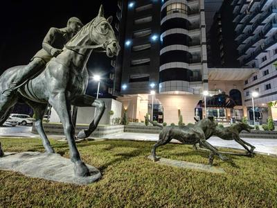 Bronzefiguren eines Reiters auf einem Pferd und zweier spielender Hunde vor beleuchtetem Hochhaus.