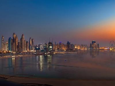 Nächtliche Skyline einer modernen Stadt mit beleuchteten Wolkenkratzern und ruhigem Wasser im Vordergrund.