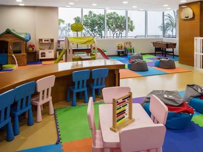 Bunter Kindergartenraum mit Tischen, Stühlen, Spielmatten und Spielgeräten vor großen Fenstern.