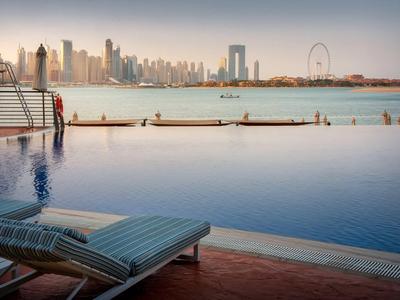 Großer Pool mit Liegestuhl, Mann am Rand und Skyline mit Wolkenkratzern im Hintergrund bei Sonnenuntergang.