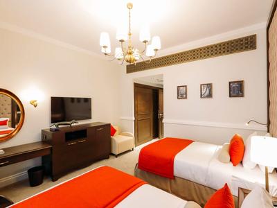 Hell eingerichtetes Zimmer mit zwei Einzelbetten, roten Decken, Tisch, Fernseher und dekorativen Bildern.