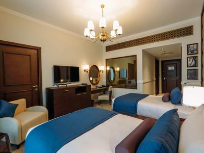 Hotelový pokoj se dvěma postelemi, televizí, židlemi a zrcadlem v teplých barvách.