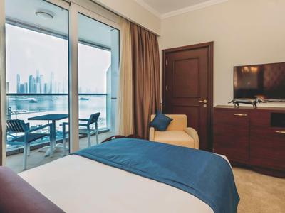 Schlafzimmer mit großem Fenster, Balkon, blauem Bettbezug, beigem Sessel und TV auf braunem Schrank.