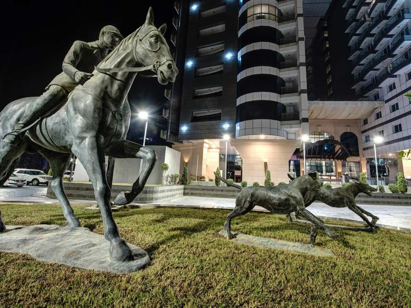 Bronzefiguren eines Reiters auf einem Pferd und zweier spielender Hunde vor beleuchtetem Hochhaus.